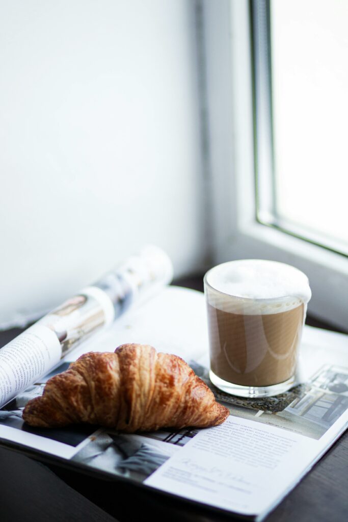 Fresh croissant and cappuccino on a magazine by a window, perfect morning setting.